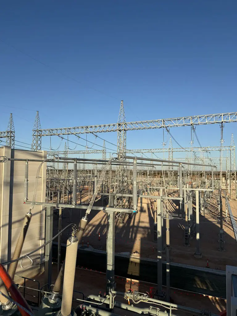 Electrical substation with numerous metal towers, power lines, and equipment under a clear blue sky. Shadows from the structures cast patterns on the ground, while ecological consultants Australia monitor activity near orange cones by a fenced area.