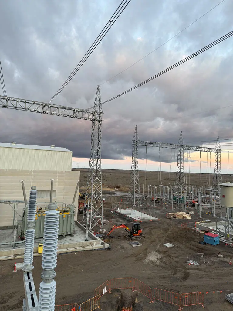 A construction site for an electrical substation with metal towers, power lines, and equipment under a cloudy sky. Ecological consultants Australia oversee as workers, vehicles, and fencing are visible on the dirt ground.