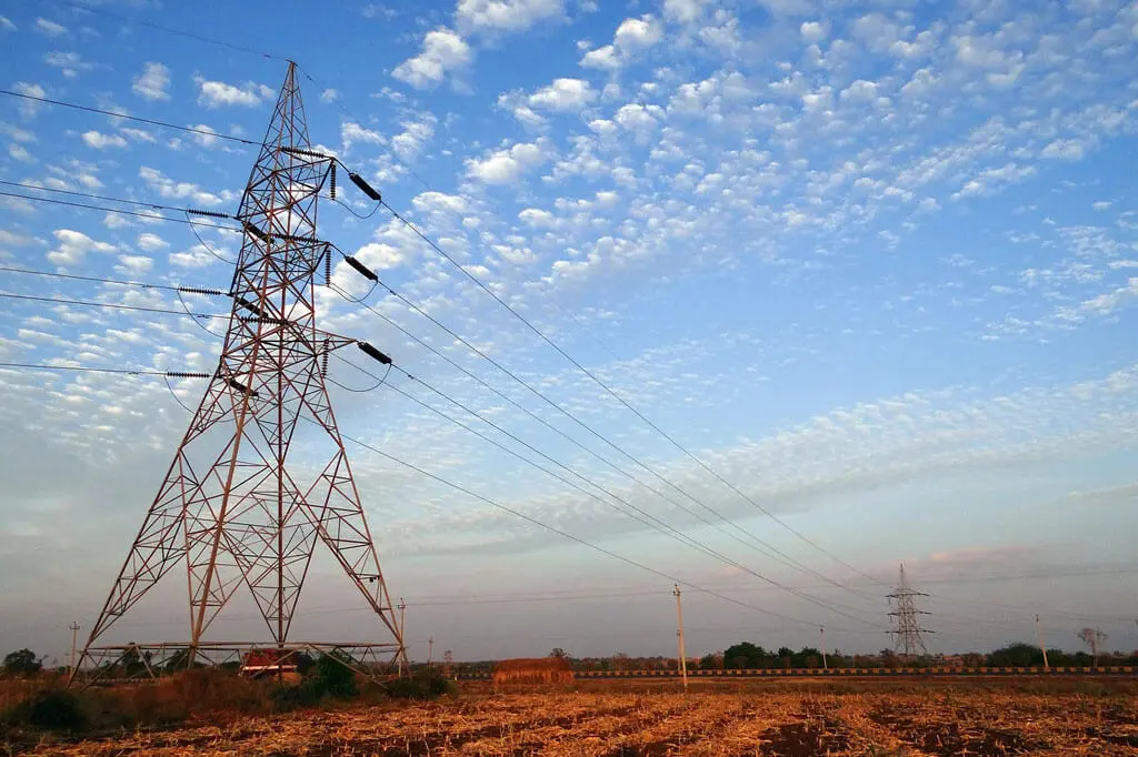 A tall electricity transmission tower stands in a dry, harvested field under a blue sky dotted with scattered clouds, as ecological consultants Australia often assess such landscapes; more towers and power lines can be seen in the distance.
